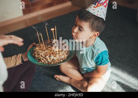 Garçon soufflant des bougies sur son gâteau d'anniversaire donné par les mains de maman tout en étant assis sur le sol pendant sa fête d'anniversaire. Banque D'Images