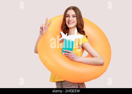 Optimiste touriste jeune fille en T-shirt jaune debout avec anneau en caoutchouc orange, tenant le passeport document et maquette d'avion, se réjouir voyage tour.Prise de vue en studio isolée sur fond gris. Banque D'Images