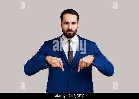 Homme barbu sérieux pointant les doigts vers le bas, exigeant une approche immédiate de lui, ayant le contrôle de la situation, portant un costume officiel.Prise de vue en studio isolée sur fond gris. Banque D'Images