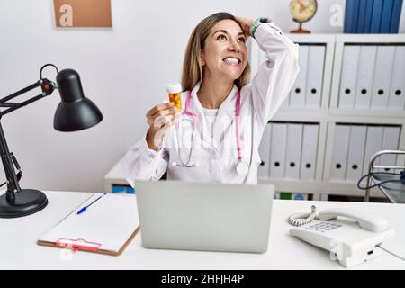 Jeune femme hispanique portant l'uniforme du médecin tenant des pilules à la clinique souriant confiant touchant les cheveux avec le geste de main vers le haut, posant attrayant et fa Banque D'Images
