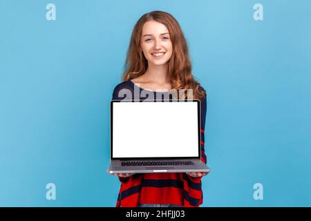 Femme gaie portant un chandail rayé style décontracté, debout montrant ordinateur portable avec écran blanc vide pour la promotion, regardant l'appareil photo avec le sourire.Studio d'intérieur isolé sur fond bleu. Banque D'Images