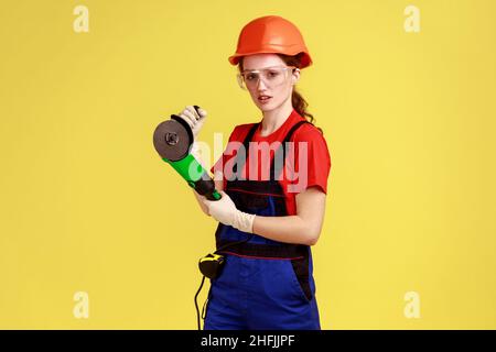 Portrait d'une femme charpentier sérieuse travaillant avec une meuleuse d'angle, ayant une expression faciale confiante, portant une combinaison et un casque de protection.Studio d'intérieur isolé sur fond jaune. Banque D'Images