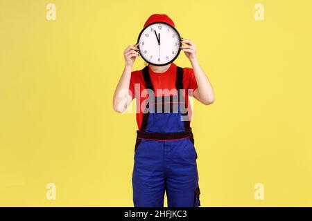 Portrait d'une travailleuse inconnue dissimulant son visage derrière une grande horloge murale, le travail sera effectué à temps, dans l'industrie des services, portant une combinaison et une casquette rouge.Studio d'intérieur isolé sur fond jaune. Banque D'Images