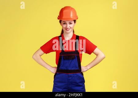 Portrait d'une femme de construction heureuse et confiante debout tenant les mains sur les hanches, regardant la caméra, portant une combinaison et un casque de protection.Studio d'intérieur isolé sur fond jaune. Banque D'Images