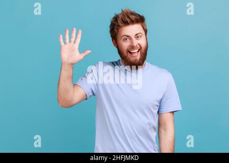 Portrait d'homme barbu positif saluant vous lever la main et en agitant, en disant salut, heureux de vous voir, regardant l'appareil photo avec un sourire en dents de cou.Studio d'intérieur isolé sur fond bleu. Banque D'Images