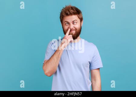 Mauvaise conduite, mauvaises manières.Portrait de l'homme barbu fou piquant le nez et collant la langue avec l'expression comique stupide, se tromper autour.Studio d'intérieur isolé sur fond bleu. Banque D'Images