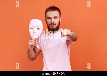 Portrait d'un homme barbu sérieux pointant du doigt vers l'appareil photo et tenant le masque blanc dans sa main, veut changer de personnalité, portant un T-shirt rose.Studio d'intérieur isolé sur fond orange. Banque D'Images