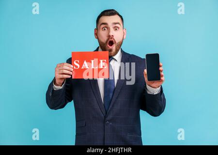 Homme d'affaires surpris avec la barbe portant le costume de style officiel tenant la carte de vente et le smartphone , choqué par l'incroyable vente en ligne.Studio d'intérieur isolé sur fond bleu. Banque D'Images