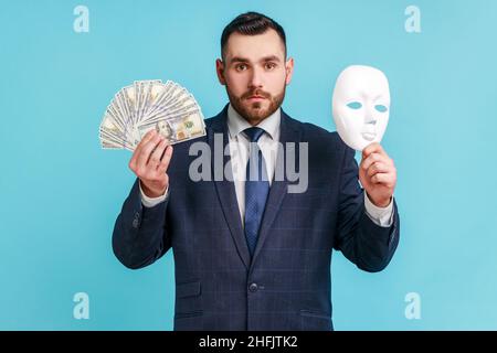 Portrait d'un homme d'affaires barbu sérieux en costume officiel tenant dans les mains des billets de banque en dollars et masque blanc, regardant l'appareil photo avec le regard strict.Studio d'intérieur isolé sur fond bleu. Banque D'Images