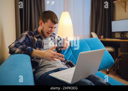 Homme assis sur le canapé et effectuant un appel vidéo.Homme barbu en colère ayant des appels vidéo avec un amant ou un collègue, à l'aide d'un ordinateur portable, regardant l'écran d'ordinateur, hurlant et gestiant, intérieur de la maison Banque D'Images