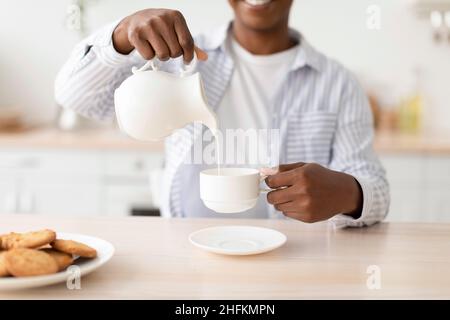 Une jeune femme noire satisfaite verse du lait dans une tasse de boisson préférée, s'assoit à table avec des biscuits à l'intérieur de la cuisine Banque D'Images