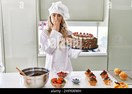 Jeune chef de pâtisserie caucasienne femme cuisant des pâtisseries et des gâteaux à la cuisine couvrant la bouche avec la main, choqué et peur de l'erreur. Surprise express Banque D'Images