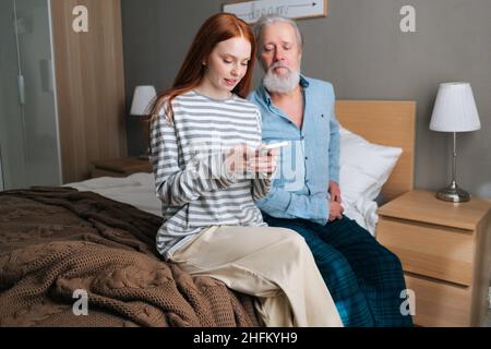 Bonne petite-fille montrant la photo sur mobile à grand-père aux cheveux gris assis sur le lit à la maison. Banque D'Images