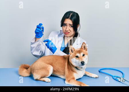 Belle femme vétérinaire hispanique mettant le vaccin au chien de chiot sceptique et nerveux, frowning bouleversé à cause du problème. Personne négative. Banque D'Images