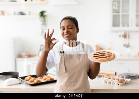 Une jolie dame noire millénaire satisfaite en tablier bénéficie de l'arôme d'une tarte fraîche et savoureuse dans un intérieur de cuisine moderne, espace libre Banque D'Images