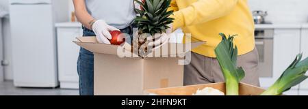 Vue rognée d'un couple en gants de latex prenant des fruits de la boîte dans la cuisine, bannière Banque D'Images