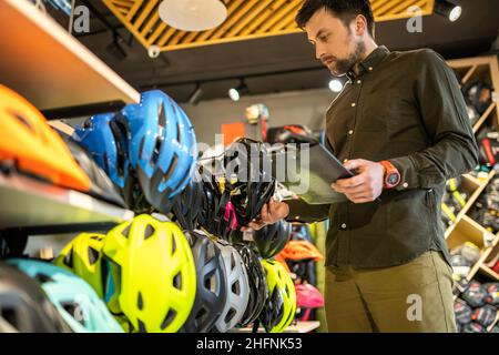 Un directeur de boutique de vélos de sexe masculin effectue un inventaire des casques de sport dans une boutique de vélos.Le propriétaire d'un magasin de sport avec un presse-papiers dans ses mains vérifie le Banque D'Images