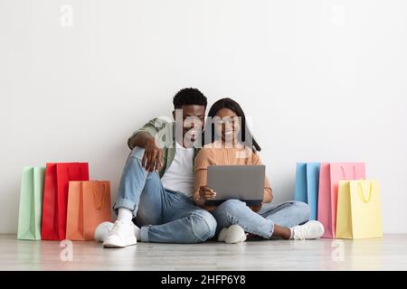 Magnifique noir amoureux homme millénaire et femme dans décontracté assis sur le sol entre les achats ensemble et en utilisant le bloc-notes, shopping en ligne, achat meubles Banque D'Images