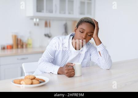 Fatigué endormi jeune femme noire attrayante avec une tasse de boisson s'assoit à la table avec des biscuits dans la cuisine scandinave intérieur Banque D'Images