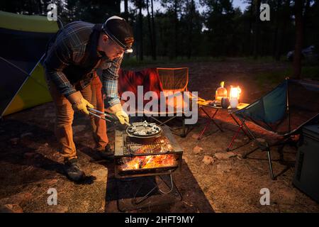 L'homme prépare un dîner de camp dans une poêle en fonte au-dessus d'un feu. Banque D'Images
