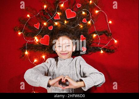 une petite fille avec une guirlande et des coeurs rouges en cheveux bouclés sourit sur un fond rouge et tient un coeur dans ses mains. Banque D'Images