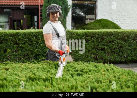 Beau caucasien homme en uniforme, défenseur du visage et gants de protection coupant des buissons trop grands sur la cour arrière.Jardinier compétent utilisant un taille-haie à essence pour le travail en extérieur. Banque D'Images