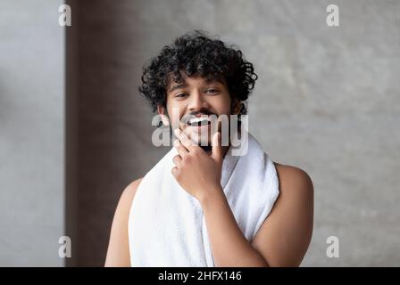 Portrait d'un Indien à barbe heureux avec une serviette posant dans la salle de bains et touchant le menton, prêt à prendre le bain Banque D'Images