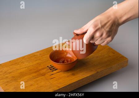 La main d'un homme verse du thé pu erh fraîchement préparé d'une théière en argile dans une tasse traditionnelle.Une tasse sur une table pour les cérémonies du thé. Banque D'Images