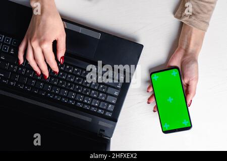 Vue de dessus des mains de femme tapant sur le clavier de l'ordinateur portable placé sur le bureau blanc avec smartphone vierge Banque D'Images