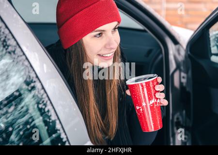 Une jeune femme heureuse avec une coupe thermo rouge est assise dans une voiture en hiver. Banque D'Images