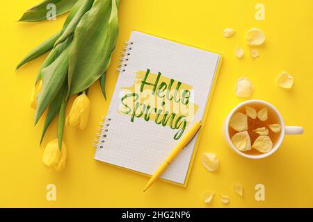 Carnet avec texte HELLO SPRING, fleurs de tulipe et tasse de thé sur fond jaune Banque D'Images