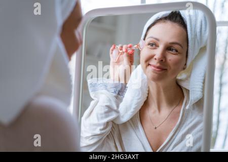 Femme utilisant le rouleau de visage.Sculpture, levage et soins anti-âge.Soins de la peau et beauté à la maison.Femme utilisant un masseur à roulettes en quartz apaisant tout en étant assise sur un canapé dans le salon et regardant dans le miroir Banque D'Images