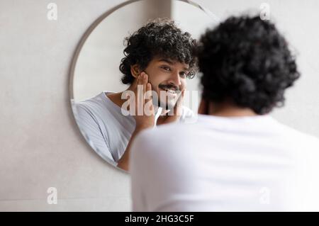 Joli barbu indien touchant le visage appliquant une crème hydratante sur le visage, debout près du miroir dans la salle de bains moderne Banque D'Images