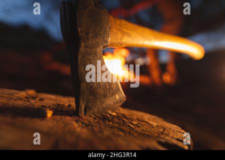 Gros plan d'une hache illuminée sur une souche d'arbre devant un feu de camp en feu de forêt Banque D'Images