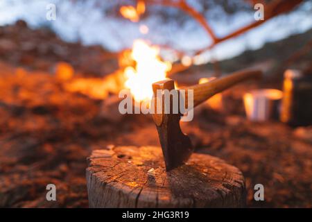 Gros plan d'une hache sur une souche d'arbre devant un feu de camp en feu de forêt au crépuscule Banque D'Images