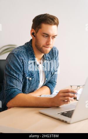 Photo verticale d'un jeune homme d'affaires sérieux dans un écouteur ayant une visioconférence en ligne sur un ordinateur portable au bureau à domicile. Banque D'Images