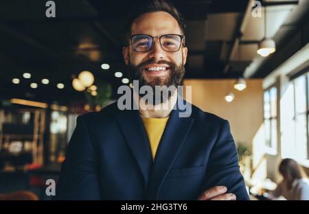 Homme d'affaires souriant à la caméra dans un espace de travail commun.Un jeune homme d'affaires heureux debout seul avec ses bras croisés.Jeune entre réussi Banque D'Images