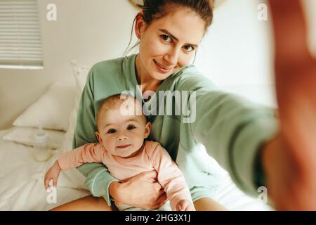 Maman blogueuse prenant un selfie avec son bébé à la maison.Maman millénaire prenant une photo tout en étant assise sur le lit avec son bébé.Maman influenceur créant con Banque D'Images