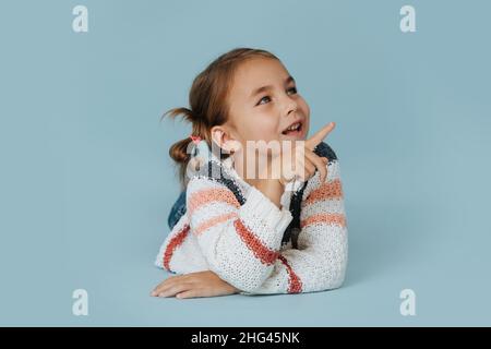 Petite fille sociale en chandail rayé couché sur le sol sur fond bleu.Parler à quelqu'un tout en articulant avec son index. Banque D'Images