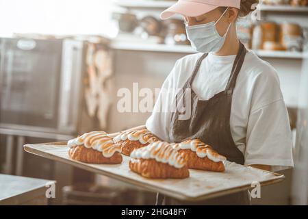 Femme dans le masque de protection tient un plateau avec des croissants savoureux décorés dans la cuisine du magasin de boulangerie Banque D'Images