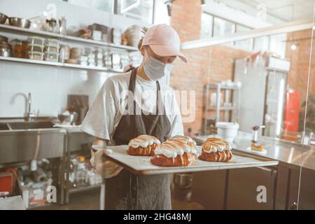 Baker dans le masque médical tient un plateau avec de beaux croissants dans la cuisine moderne Banque D'Images