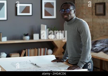 Jeune architecte contemporain en lunettes et pull gris regardant la caméra tout en se tenant debout à table avec plan Banque D'Images