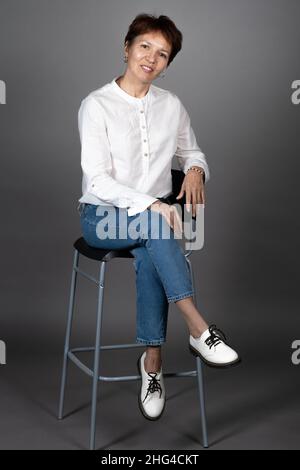 Portrait d'une belle femme mûre posant sur une chaise en chemisier et jeans sur fond gris Banque D'Images