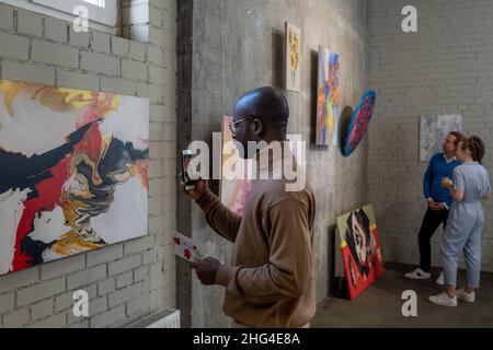 Jeune Africain visiteur de la galerie d'art prenant photo de la peinture sur le mur tout en appréciant l'exposition des artistes modernes Banque D'Images