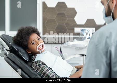 Petit garçon avec un rétracteur médical sur la bouche vous regardant dans un fauteuil dans les cliniques dentaires avant la procédure de blanchiment Banque D'Images