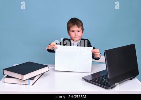 Un garçon dans un costume d'école avec une feuille de papier de A4 à un ordinateur pendant l'apprentissage à distance, un espace de copie sur un fond bleu studio Banque D'Images
