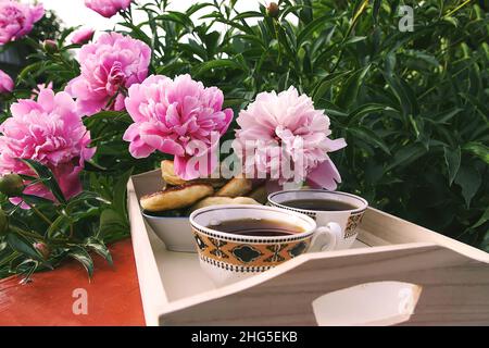 Thé de style campagnard dans le jardin d'été du village.Deux tasses de thé noir chaud sur plateau en bois et fleurs de pivoine rose en plein soleil. Banque D'Images