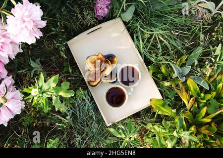 Thé de style campagnard dans le jardin d'été du village.Deux tasses de thé noir chaud et crêpes faites maison sur plateau en bois et fleurs de pivoine rose i Banque D'Images