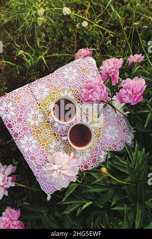 Thé de style campagnard dans le jardin d'été du village.Deux tasses de thé noir chaud sur une nappe de dentelle vintage crochetée et fleurs de pivoine au soleil Banque D'Images