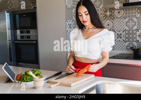 Femme élégante épluchant des carottes fraîches près de l'assiette avec des poivrons et un comprimé avec une recette tout en cuisinant un déjeuner sain dans la cuisine à la maison Banque D'Images